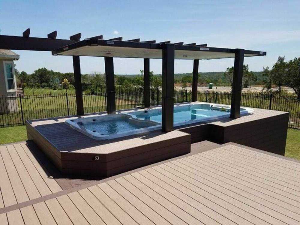 American Whirlpool porch spa with raisable cover, surrounded by a wooden deck and pergola, set in a landscaped outdoor area.