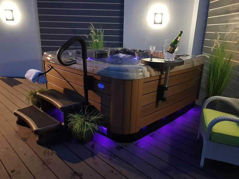 American Whirlpool spa with champagne, illuminated steps, and surrounding plants on a wooden deck, highlighting relaxation and luxury in outdoor settings.