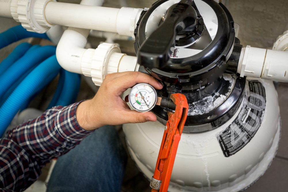Hand adjusting a pressure gauge on a pool filter system, with blue hoses and plumbing visible, related to pool maintenance and service at Leisure Pools & Spas.