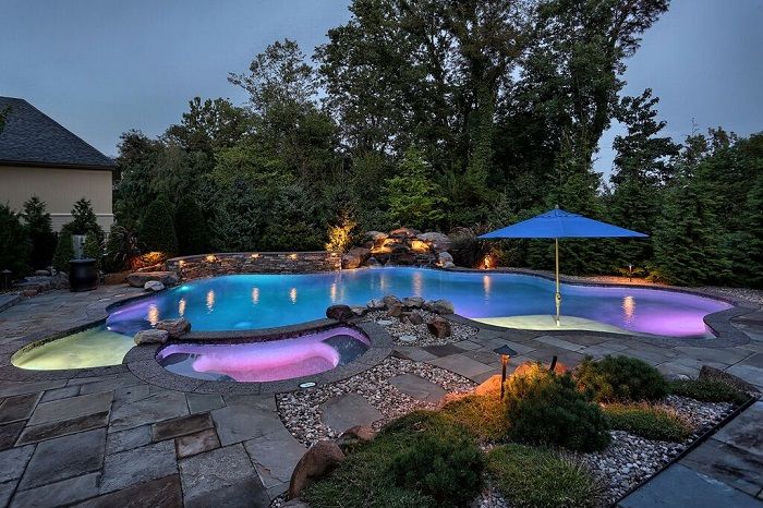 Illuminated swimming pool with vibrant lighting, surrounded by stone patio, lush greenery, and a blue umbrella, showcasing a serene outdoor environment ideal for relaxation and pool maintenance services.