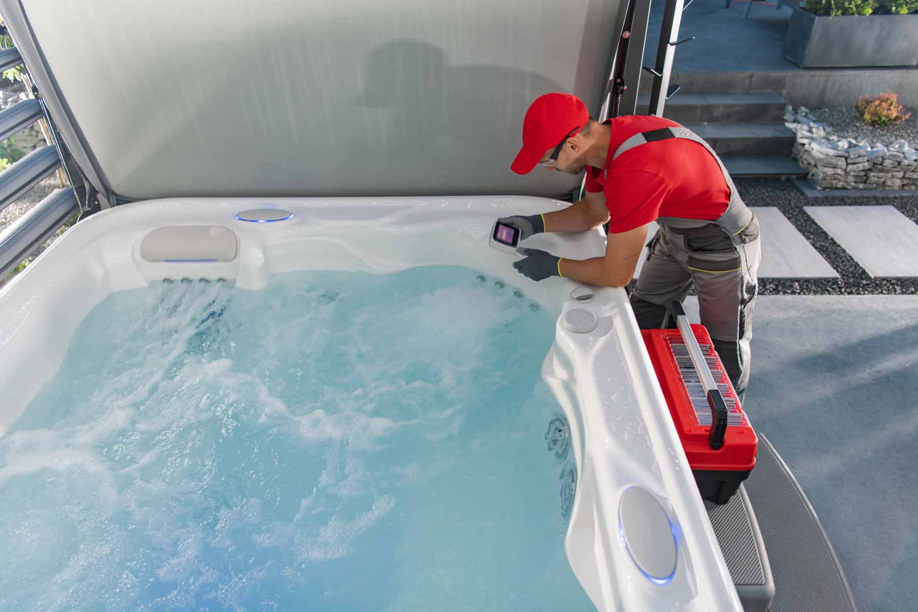 Technician performing maintenance on a hot tub, using a digital device for water testing, with tools and equipment visible nearby.