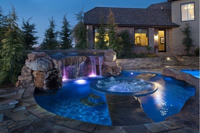 Luxury spa and pool with waterfall feature, illuminated by colorful lights, surrounded by landscaping and a home in the background, representing professional pool and spa maintenance services.