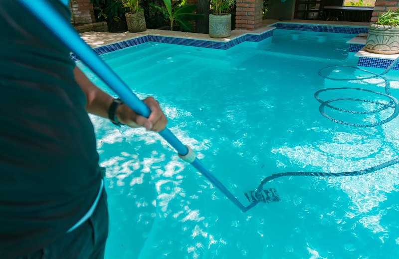 Person using a pool vacuum to clean a swimming pool, showcasing routine maintenance services for optimal water clarity and pool performance.