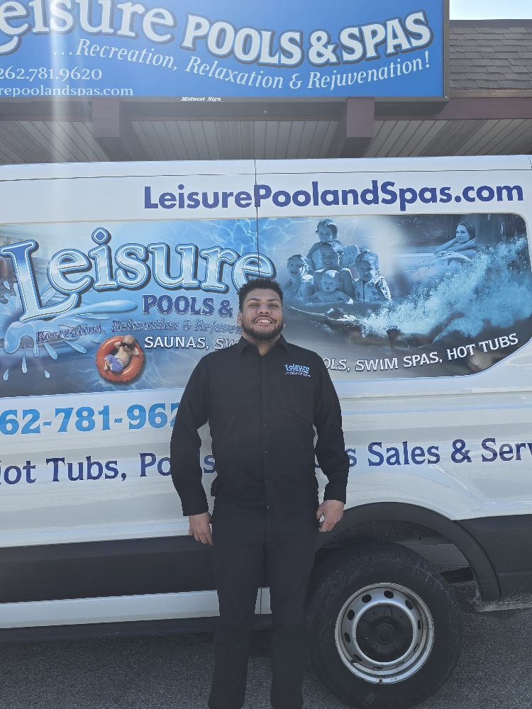 Man standing in front of a Leisure Pools & Spas vehicle, wearing a black shirt with the company logo, smiling, and showcasing the business's branding and services related to pools and hot tubs.