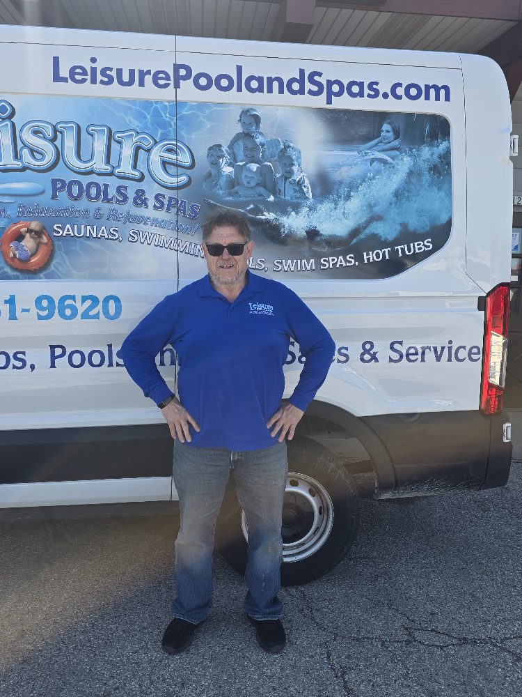 Man in blue Leisure Pools & Spas polo shirt standing confidently in front of service van featuring company branding and pool-related imagery, emphasizing expertise in pool and hot tub services.