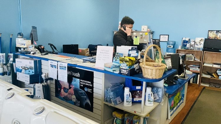 Store interior of Leisure Pools & Spas featuring a customer service representative on the phone, displaying pool and spa supplies, accessories, and promotional materials on a blue counter.