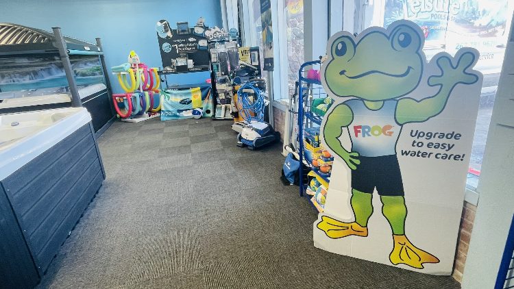 Store interior featuring a cartoon frog mascot promoting water care, alongside pool supplies and accessories, including hot tubs and cleaning equipment, at Leisure Pools & Spas.