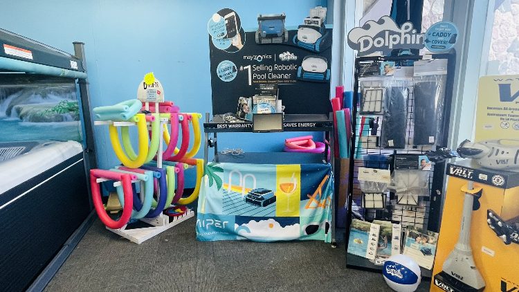 Colorful pool accessories and cleaning equipment displayed in a store, featuring a prominent robotic pool cleaner, colorful pool noodles, and various pool maintenance products, highlighting leisure and outdoor enjoyment.